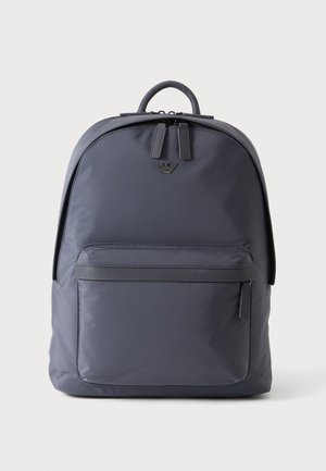 Sac à dos gris foncé avec une poche avant zippée, une fermeture double zip en haut, et un petit logo en métal au centre supérieur avant.