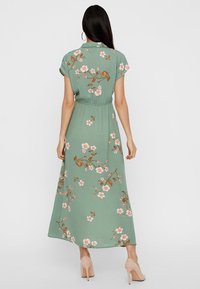 Robe longue verte à manches courtes, motif floral en rose et marron, taille élastique et design fluide, associée à des talons nus.