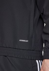 Zwarte sportjack met een gladde textuur, elastische tailleband en wit "AEROREADY" logo aan de onderzijde.