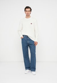 Weißer gestrickter Pullover mit Rundhalsausschnitt und Logopatch, kombiniert mit blauen gemusterten weitgeschnittenen Jeans und weißen Sneakers.