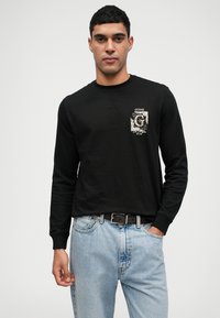 Camisola preta de manga longa com uma impressão gráfica na frente e decote redondo, combinada com jeans de ganga azul claro e um cinto preto.