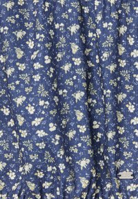 Blau gemusterter Stoff mit weißen und gelben Blumen, strukturierte Oberfläche mit leichtem Rippeneffekt und einem silbernen Markenschild an der Seite.