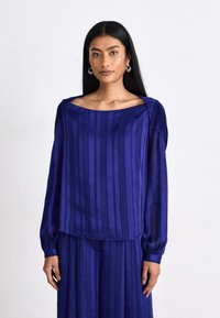 Royal blå bluse med vertikale striper og puffede ermer. Har en bred, firkantet utringning og myk, flytende stoff.