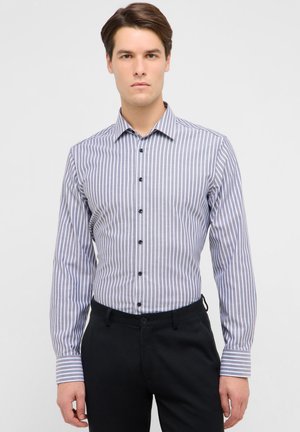 Jeune homme portant une chemise ajustée à manches longues à rayures verticales boutonnée, rentrée dans un pantalon noir, debout devant un fond uni.