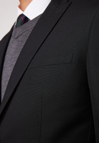 Giacca da uomo nera con finitura strutturata, dotata di taschino al petto e revers affilati, indossata sopra un maglione grigio a scollo a V.