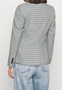 Blazer a scacchi in luce blu, beige e nero; vestibilità su misura con maniche lunghe e dettagli con bottoni neri sui polsini. Indossato con jeans in denim chiaro.