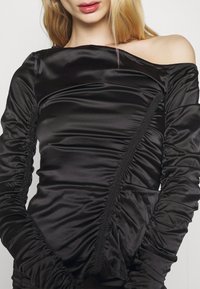 Eine Frau trägt ein gerafftes, schulterfreies schwarzes Satintop mit langen Ärmeln, das ihren Hals, Schlüsselbein und einen Teil ihrer Lippen zeigt.