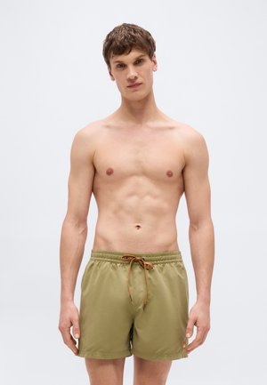 Mężczyzna bez koszulki z krótkimi brązowymi włosami, ubrany w oliwkowo-zielone kąpielówki z pomarańczowym sznurkiem, stojący na tle jednolitego jasnego tła.