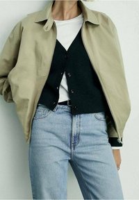 Personne portant une veste zip beige clair par-dessus un gilet noir boutonné et une chemise blanche, associée à un jean taille haute bleu clair.