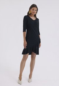 Czarna sukienka w białe kropki z rękawami do trzech czwartych, falbaną u dołu i dekoltem w kształcie litery V. Noszona z beżowymi butami na szpilce i złotymi kolczykami w kształcie kółek.