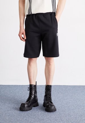 Osoba nosząca czarne krótkie spodenki Adidas oraz czarne wojskowe buty na sznurowadła stoi na szarym dywanie.