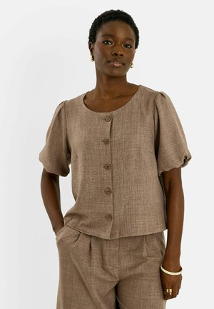 Mujer con una blusa marrón de manga corta texturizada con botones y pantalones de cintura alta a juego, con pendientes y pulseras doradas, posando con una mano en el bolsillo.