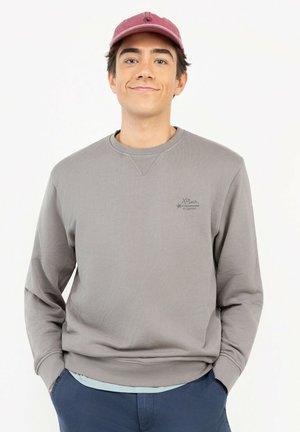 Joven con gorra rosa apagado, sudadera gris con logo y pantalones azules, de pie con las manos en los bolsillos sobre fondo blanco.