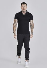 Camisa polo negra con cremallera combinada con pantalones jogger negros y zapatillas blancas. Incluye tatuajes y una pose casual sobre un fondo neutro.