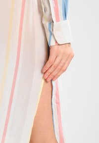 Robe chemise rayée en tissu transparent présentant des rayures verticales dans des couleurs pastel (rose, bleu, jaune) avec une fente sur le côté et des poignets boutonnés.