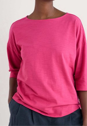Femme portant une chemise ample rose vif à manches mi-longues et un pantalon bleu foncé, mains dans les poches, sur un fond uni.