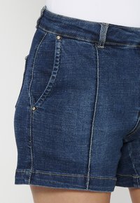 Jeans kratke hlače v temno modri barvi, s klasičnim krojem, dvema stranskima žepoma s dekorativnim šivom in kontrastnimi zlatimi dodatki.