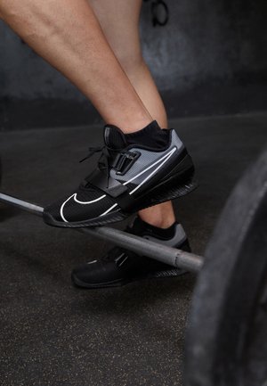 Athlète portant des chaussures d'haltérophilie noires et grises marchant sur une barre d'haltères sur le sol d'une salle de sport.