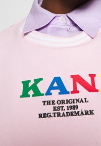 Sudadera rosa con escote redondo, que presenta un texto en negrita de múltiples colores "KAN" y "THE ORIGINAL EST. 1989" en negro, superpuesto a una camisa de cuello ligera de color púrpura.