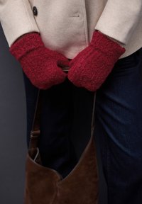 Rode, gestructureerde wanten gecombineerd met een beige jas, die een bruine suède schoudertas vasthoudt, gedragen met donkere jeans.