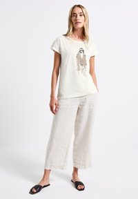 T-shirt crème clair avec une illustration graphique, associé à un pantalon en lin beige ample et des sandales à glissière noires ; coupe décontractée et tissu doux.