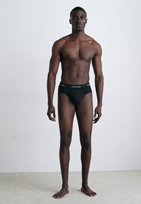 Schwarze Baumwollslips mit einem gebrandeten Bund, taillierten Design und glatter Textur, getragen von einem Modell vor einem hellen Hintergrund.