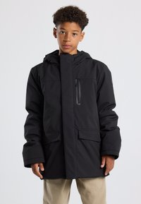 Musta huppari, jossa on täysi etuvetoketju, kaksi sivutaskua ja pieni rintatasku vetoketjulla; materiaalissa on sileä, vettä hylkivä pinta.