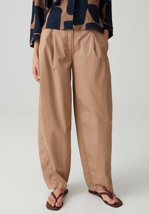 Vrouw draagt losse beige broek gecombineerd met een zwart-bruin patroon overhemd en bruine slippers.