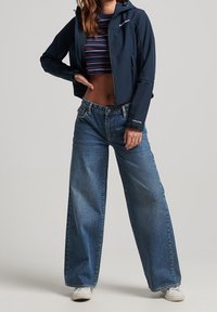 Marinblå zip-jacka med huva, kombinerad med randig kort topp och lösa, urtvättade blå jeans med vida ben. Vita sneakers kompletterar outfiten.