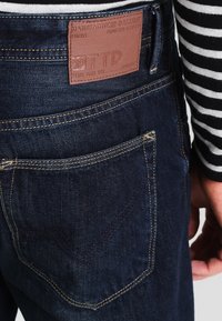 Vaqueros de mezclilla azul oscuro con una textura suave, que cuentan con un parche de cuero marrón, costuras en contraste y un bolsillo trasero con una costura diagonal.