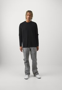 Suéter negro de punto, manga larga, con cuello redondo. Jeans gris claro desgastados con bolsillos. Zapatillas blancas completan el atuendo.