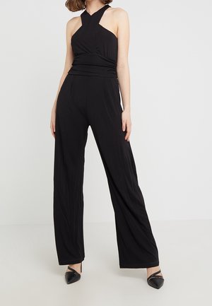 Mujer con un mono negro de tirantes estilo halter sin mangas, con pantalones anchos y tacones negros de punta puntiaguda sobre un fondo liso.