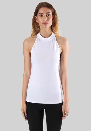 Femme aux cheveux ondulés mi-longs, portant un haut blanc sans manches à col halter et un pantalon noir, debout devant un fond gris uni.