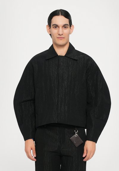 Chaqueta negra corta con una superficie texturizada, cuello puntiagudo y mangas anchas. Cuenta con una pequeña etiqueta removible en la cintura.