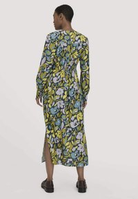 Langes Kleid mit Blumenmuster in grünen, blauen und gelben Farbtönen, mit langen Ärmeln, einem gerafften Rücken und seitlichen Schlitzen. Getragen mit dunklen Schuhen.