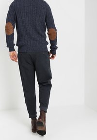 Maglione a coste blu navy con toppe marroni sui gomiti, abbinato a pantaloni scuri e affusolati e stivaletti marroni. La texture appare lavorata a maglia e liscia.