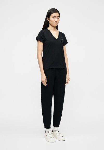 Νεαρή γυναίκα που στέκεται, φορώντας μαύρο T-shirt με V-λαιμόκοψη, μαύρο παντελόνι jogger, λευ κές κάλτσες και λευκά αθλητικά παπούτσια, μπροστά σε απλό φόντο.