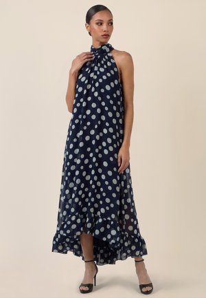 Mujer con un vestido azul marino sin mangas con lunares blancos, escote halter, dobladillo con volantes y sandalias negras de tacón alto.