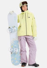 Donna che indossa una giacca gialla, pantaloni da neve rosa, guanti bianchi e casco, mentre tiene una tavola da snowboard con un design floreale blu.
