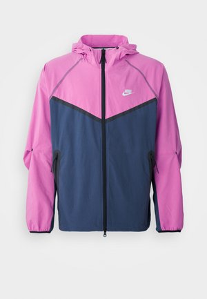 Nike vindjacka med en rosa överdel och marinblå nederdel, försedd med en framdragbar dragkedja, huva och sidofickor.