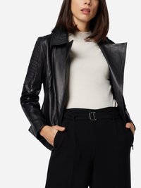 Schwarze Lederjacke mit gesteppten Schultern, Reverskragen und Reißverschluss; getragen über einem hellen Oberteil und schwarzen hoch taillierten Hosen.