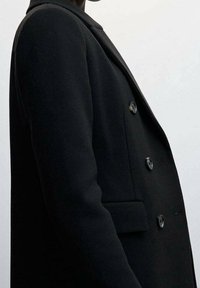 Een persoon in een zwart dubbelzijdig jasje met drie zichtbare knopen, staand tegen een effen lichte achtergrond.