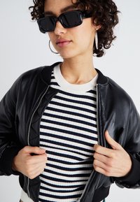 Black leather bomber jacket, ribbed white and navy striped shirt, and rectangular black sunglasses. Notable silver zipper and hoop earrings.