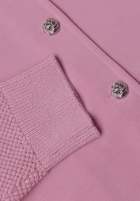 Gros plan sur une manche tricotée rose et un tissu assorti avec deux boutons décoratifs argentés disposés en diagonale.