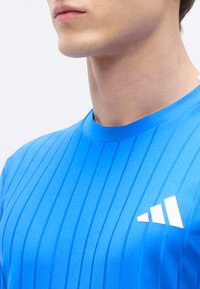 Chemise de sport bleue en tissu lisse, présentant des rayures verticales et un logo blanc sur la partie gauche de la poitrine.