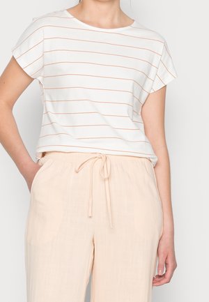 Frau trägt ein weißes Kurzarmshirt mit dünnen horizontalen orangen Streifen und hellbeige Hosen mit Kordelzug, Hände seitlich am Körper.