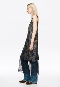 Vestido de cuadros negro y marrón con dobladillo de encaje, que presenta tirantes finos y un corte suelto, superpuesto a unos pantalones de denim anchos con acentos de encaje.