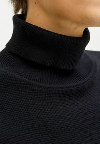 Maglione a collo alto nero a costine che mostra un tessuto texturizzato e un colletto aderente. I dettagli delle cuciture sono visibili, evidenziando il design a maglia.
