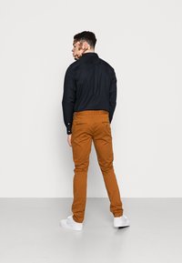 Schwarzes Button-Up-Hemd, braune Chinos mit Taschen und weiße Sneakers; von hinten vor einem schlichten Hintergrund betrachtet.