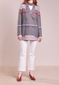 Blazer doppiopetto a scacchi grigi con strisce rosse e blu, abbinato a pantaloni a zampa d'elefante bianchi e scarpe con tacco rosse.
